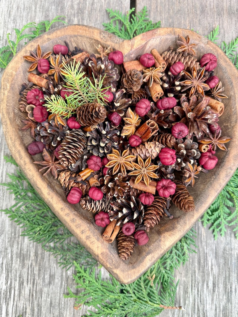 May include: A heart-shaped wooden bowl filled with pine cones, star anise, cinnamon sticks, and small red decorative items. Green sprigs of pine and cedar surround the bowl, creating a rustic, natural arrangement.