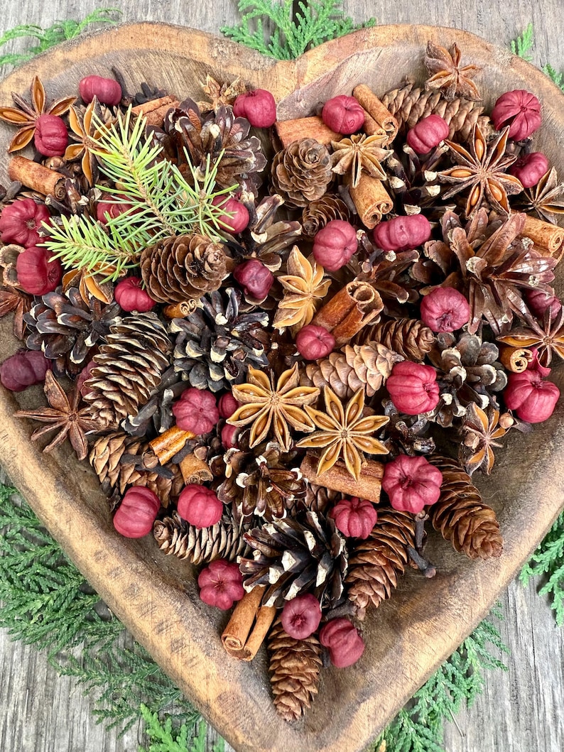 May include: A heart-shaped wooden bowl filled with a decorative arrangement of pine cones, star anise, cinnamon sticks, and small red decorative pumpkins. Green sprigs add a touch of freshness to the rustic display.