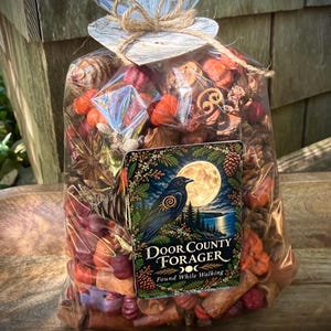 May include: A clear plastic bag filled with assorted dried botanicals, including pinecones, orange pumpkins, and cinnamon sticks. The bag is tied with twine and a heart-shaped tag. A label features a black crow, a full moon, and the text Door County Forager.
