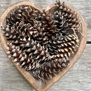 May include: A heart-shaped wooden bowl filled with brown pine cones. The bowl is sitting on a wooden surface.