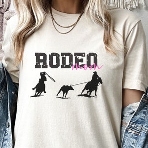 May include: A white t-shirt with the text "RODEO mom" in black with a pink "mom". The shirt features a silhouette of a cowboy roping a calf.