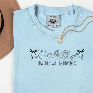 May include: A light blue t-shirt with the text "COWGIRLS WILL BE COWGIRLS" and a graphic of a horse, hat, boots, and a cowgirl. A gold necklace with a star pendant is draped across the shirt. The shirt has a Comfort Colors tag.