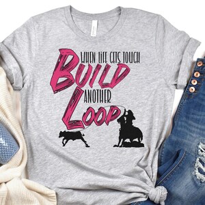 May include: Gray t-shirt with a pink graphic that says "When life gets tough build another loop". The graphic also includes a silhouette of a cowboy roping a calf.