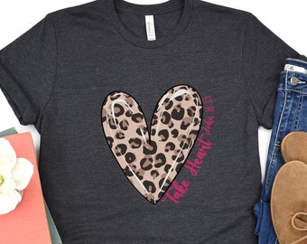 May include: A dark grey t-shirt with a pink leopard print heart and the text "Take Heart John 16:33".