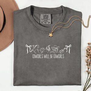 May include: A gray Comfort Colors t-shirt with the text "COWGIRLS WILL BE COWGIRLS" in white. The design includes a horse, cowboy hat, boots, and a cowgirl. A gold star pendant necklace is draped across the shirt.