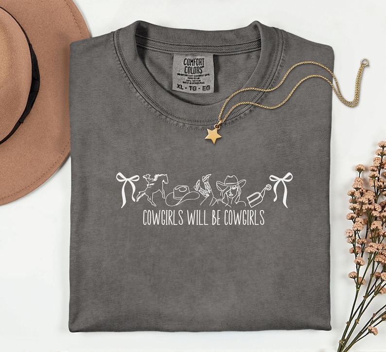 May include: A gray Comfort Colors t-shirt with the text "COWGIRLS WILL BE COWGIRLS" in white. The design includes a horse, cowboy hat, boots, and a cowgirl. A gold star pendant necklace is draped across the shirt.