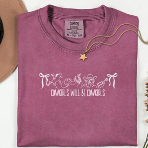 May include: A dusty rose Comfort Colors t-shirt with the text "COWGIRLS WILL BE COWGIRLS" in white. The design includes a horse, cowboy hat, and other western-themed graphics. A gold star pendant necklace rests on the shirt.