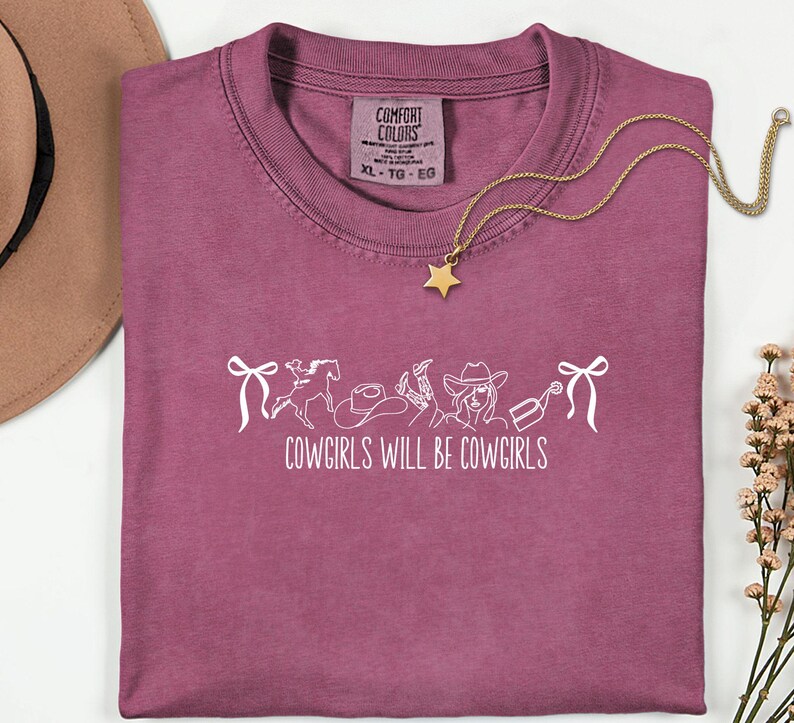 May include: A dusty rose Comfort Colors t-shirt with the text "COWGIRLS WILL BE COWGIRLS" in white. The design includes a horse, cowboy hat, and other western-themed graphics. A gold star pendant necklace rests on the shirt.