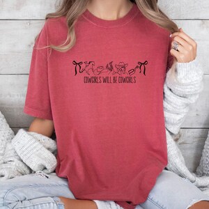 May include: A red-orange t-shirt with the text "COWGIRLS WILL BE COWGIRLS" in black. The shirt features a line drawing of a bow, a horse, a hat, a boot, and a bottle. The shirt is made of a soft material.