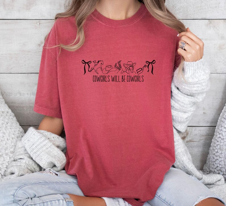 May include: A red-orange t-shirt with the text "COWGIRLS WILL BE COWGIRLS" in black. The shirt features a line drawing of a bow, a horse, a hat, a boot, and a bottle. The shirt is made of a soft material.