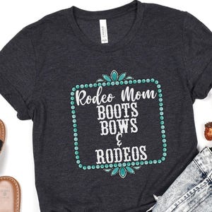 May include: A dark gray t-shirt with a turquoise and white design. The design features a square frame with turquoise jewels and the text "Rodeo Mom Boots Bows & Rodeos".