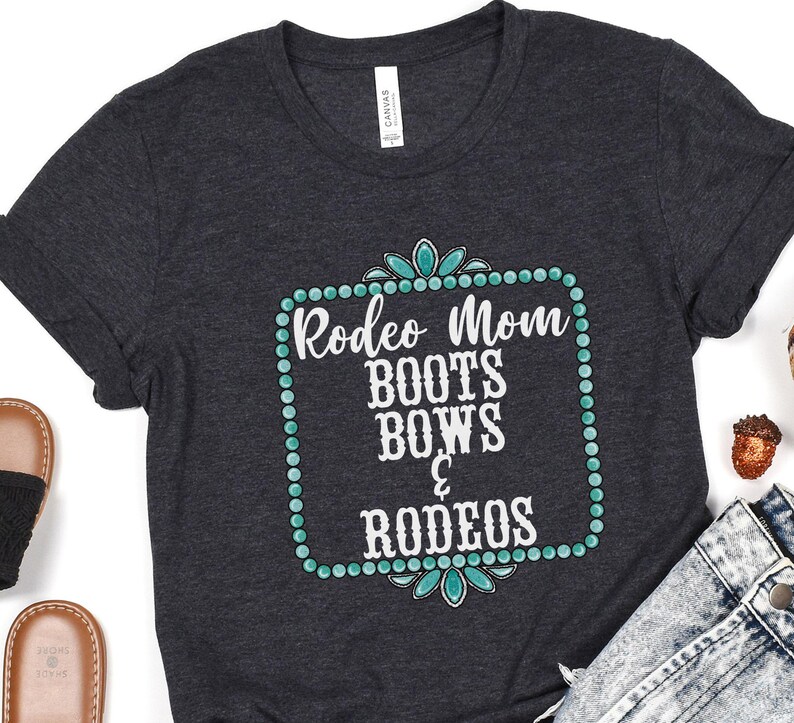 May include: A dark gray t-shirt with a turquoise and white design. The design features a square frame with turquoise jewels and the text "Rodeo Mom Boots Bows & Rodeos".