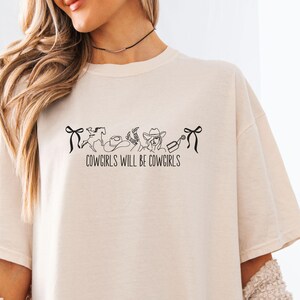 May include: Cream-colored t-shirt with the text "COWGIRLS WILL BE COWGIRLS" in black. The design includes line art of a horse, cowboy hat, and other western-themed elements. The shirt has a relaxed fit.