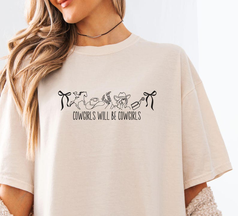 May include: Cream-colored t-shirt with the text "COWGIRLS WILL BE COWGIRLS" in black. The design includes line art of a horse, cowboy hat, and other western-themed elements. The shirt has a relaxed fit.