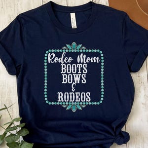 May include: A navy blue t-shirt with a white graphic that says "Rodeo Mom Boots Bows & Rodeos" in a turquoise and white frame.