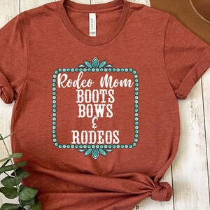 May include: A rust-colored t-shirt with a turquoise and white design that says "Rodeo Mom Boots Bows & Rodeos".