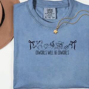 May include: A light blue Comfort Colors t-shirt with the text "COWGIRLS WILL BE COWGIRLS" and a black graphic of cowboy-themed images. A gold star pendant necklace is draped over the shirt. The shirt is size XL.