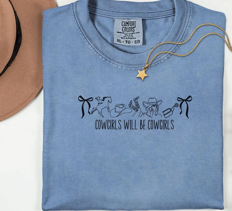 May include: A light blue Comfort Colors t-shirt with the text "COWGIRLS WILL BE COWGIRLS" and a black graphic of cowboy-themed images. A gold star pendant necklace is draped over the shirt. The shirt is size XL.