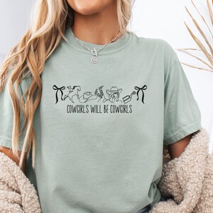 May include: A light green t-shirt with the text "COWGIRLS WILL BE COWGIRLS" in black. The graphic includes a horse, cowboy hat, boots, and a cowgirl. The shirt has short sleeves and a relaxed fit.