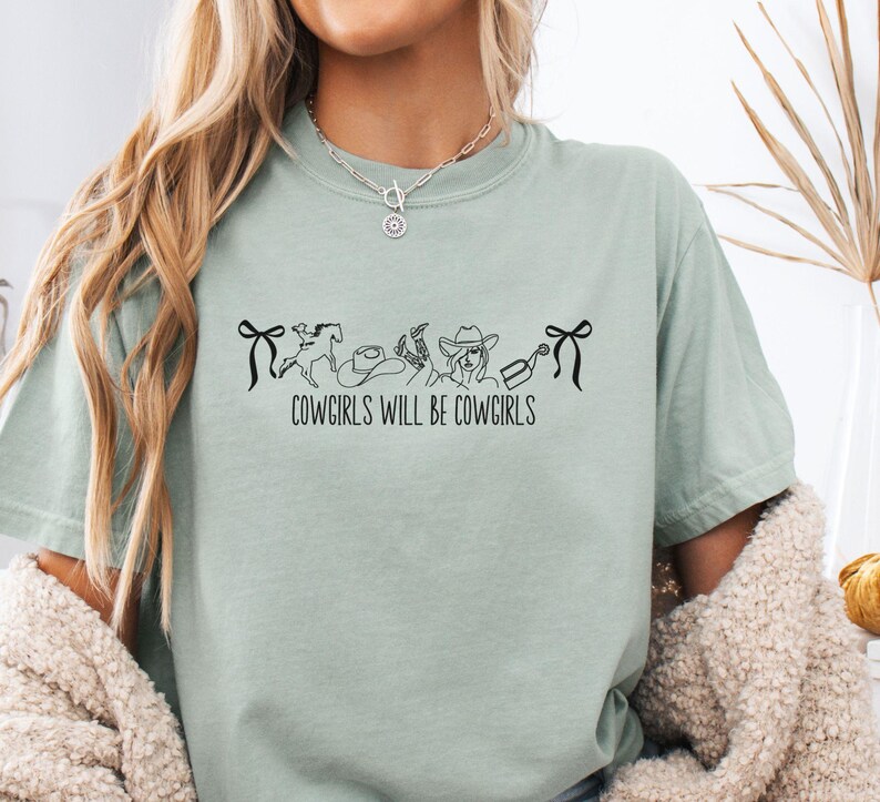May include: A light green t-shirt with the text "COWGIRLS WILL BE COWGIRLS" in black. The graphic includes a horse, cowboy hat, boots, and a cowgirl. The shirt has short sleeves and a relaxed fit.