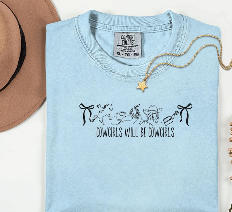 May include: A light blue t-shirt with the text "COWGIRLS WILL BE COWGIRLS" and a graphic of a horse, hat, boots, and a cowgirl. A gold necklace with a star pendant is draped across the shirt. The shirt has a Comfort Colors tag.