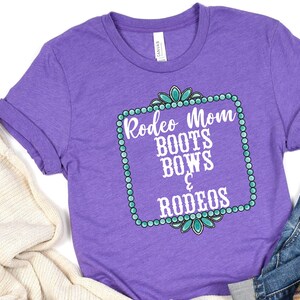May include: Purple t-shirt with a white graphic that says "Rodeo Mom Boots Bows & Rodeos" in a turquoise and white frame.