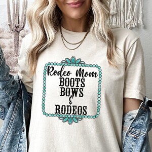 May include: A white t-shirt with a turquoise and black design that says "Rodeo Mom Boots Bows & Rodeos".