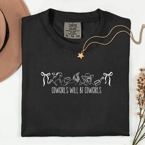 May include: Black t-shirt with a white graphic of cowgirl-themed illustrations and the text "COWGIRLS WILL BE COWGIRLS". A gold necklace with a star pendant is draped on the shirt. A brown hat is visible in the background.