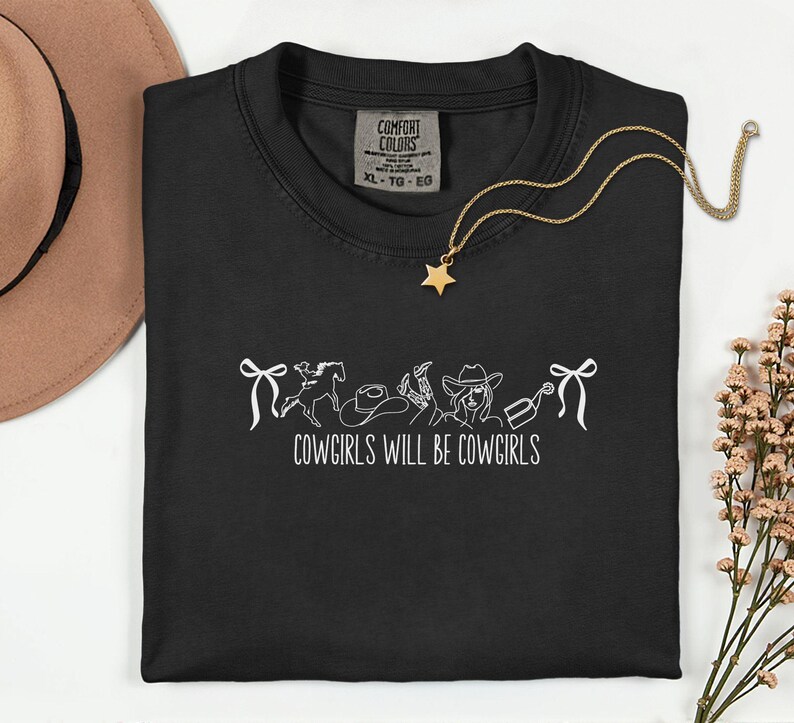 May include: Black t-shirt with a white graphic of cowgirl-themed illustrations and the text "COWGIRLS WILL BE COWGIRLS". A gold necklace with a star pendant is draped on the shirt. A brown hat is visible in the background.