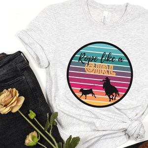 May include: A white t-shirt with a retro-style graphic of a cowgirl roping a calf. The graphic is in a circle with a rainbow-colored background and the text "Rope like a GIRL" in black and orange letters.