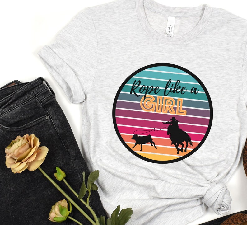 May include: A white t-shirt with a retro-style graphic of a cowgirl roping a calf. The graphic is in a circle with a rainbow-colored background and the text "Rope like a GIRL" in black and orange letters.