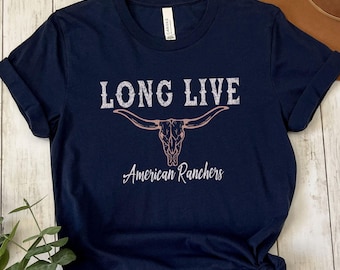 May include: Navy blue t-shirt with the text "LONG LIVE" above a steer skull graphic and the words "American Ranchers" in a script font. The shirt is made of a soft material and has rolled-up sleeves.