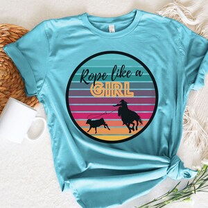 May include: A turquoise blue t-shirt with a graphic of a cowgirl roping a calf in front of a sunset. The graphic is in a circle with the text "Rope like a GIRL" in a retro font.