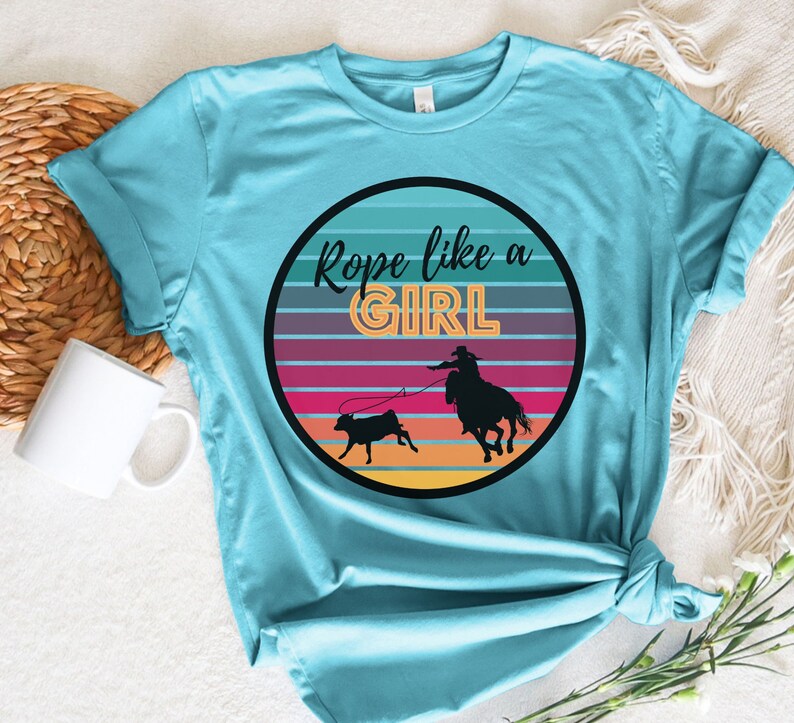 May include: A turquoise blue t-shirt with a graphic of a cowgirl roping a calf in front of a sunset. The graphic is in a circle with the text "Rope like a GIRL" in a retro font.