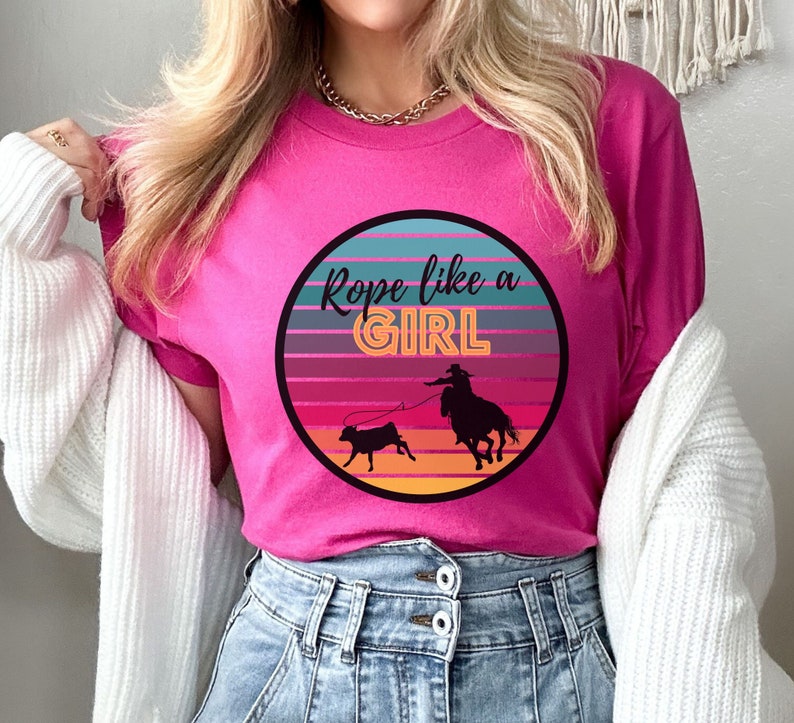 May include: Pink t-shirt with a retro-style graphic of a cowgirl roping a calf. The graphic says "Rope like a GIRL" in a bold, orange font.