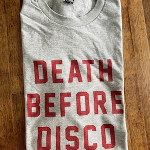 May include: A gray t-shirt with the words "DEATH BEFORE DISCO" printed in red block letters. The shirt is folded and lies on a wooden surface. The shirt has a crew neck and a small tag at the neckline.