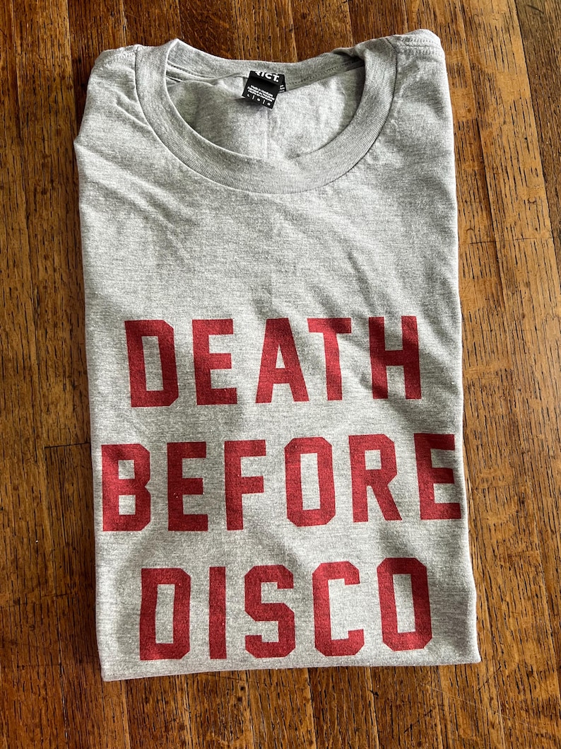 May include: A gray t-shirt with the words "DEATH BEFORE DISCO" printed in red block letters. The shirt is folded and lies on a wooden surface. The shirt has a crew neck and a small tag at the neckline.