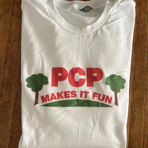 May include: White t-shirt with the text "PCP MAKES IT FUN" in red and green. The design includes two green trees on either side of the text, set against a green horizontal stripe.