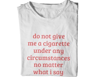 May include: White t-shirt with the red text: "do not give me a cigarette under any circumstances no matter what i say". The shirt is folded and has a crew neck.