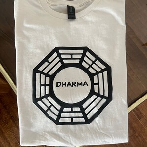 May include: White t-shirt with a black Dharma Initiative logo printed on the front. The logo is a stylized octagon with the word "DHARMA" in the center.