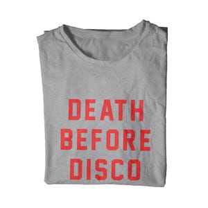 May include: A folded, light gray t-shirt with the words "DEATH BEFORE DISCO" printed in bold, red capital letters. The shirt is made of a soft material and is displayed on a white background.