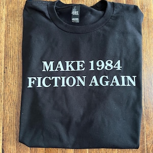 May include: Black t-shirt with the text "MAKE 1984 FICTION AGAIN" in white block letters. The shirt is folded and lies on a wooden surface. The shirt has a crew neck and short sleeves.