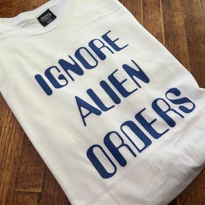 May include: White t-shirt with blue text that reads "Ignore Alien Orders".