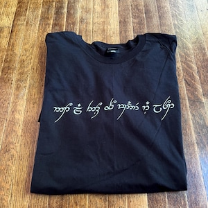 May include: Black t-shirt with a white inscription in Elvish script that reads "Ash nazg durbatulûk, ash nazg gimbatul, ash nazg thrakatulûk, agh burzum-ishi krimpatul."