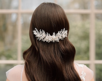May include: A silver and pearl bridal hair comb with white floral accents is set in dark brown wavy hair. The comb is worn at the back of the head, above a white lace wedding dress.