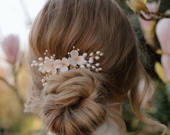 May include: A gold-toned hair comb adorned with white flowers and faux pearls. The comb is set in a woman's updo hairstyle, with a white veil visible. The background features soft-focus pink flowers and greenery, suggesting a wedding or special occasion.