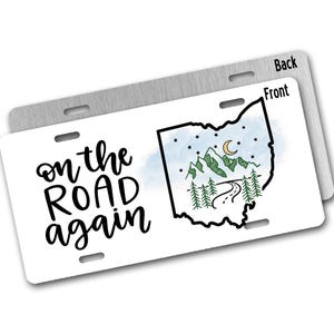 May include: White license plate with the text "on the road again" and a black outline of the state of Ohio with a watercolor background of blue sky, mountains, trees, and a crescent moon.