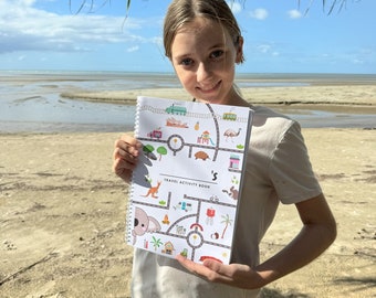 Może przedstawiać: Młoda osoba trzymająca białą książkę z grami podróżniczymi z kolorową ilustracją australijskich zwierząt i zabytków. Książka ma tytuł "Travel Activity Book" i słowo "'s" na końcu. Osoba stoi na piaszczystej plaży z błękitnym niebem i palmami w tle.