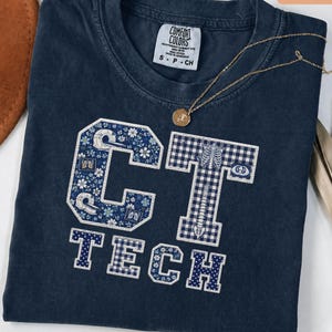 May include: A dark blue t-shirt with a crew neckline and a Comfort Colors label. The front features large block letters spelling CT TECH. The letters are filled with floral, gingham, and polka dot patterns, along with medical imaging graphics.