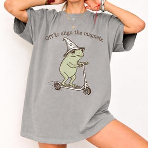 Pode incluir: Camiseta cinza com o texto "Off to align the magnets." Um sapo de desenho animado usando um chapéu de mago anda de scooter. O sapo é verde claro e a scooter é prateada.